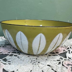 May include: A round, olive-green bowl with a black rim and white leaf-shaped designs. The bowl is made of metal and sits on a white and pink lace doily. The interior of the bowl is a lighter shade of green.
