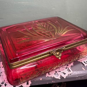 Puede incluir: Una joyero rectangular de cristal rojo rubí con ribete y cierre de metal dorado. La tapa presenta un diseño floral en relieve. La caja está sobre un tapete de encaje rosa.