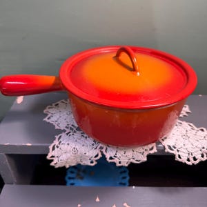 May include: A red and orange enameled saucepan with a lid and a single handle. The pot is sitting on a white lace doily. The handle and rim are red, and the body of the pot fades from orange to red.