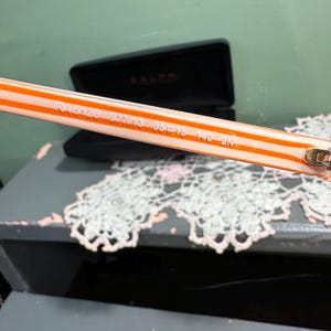Puede incluir: Primer plano de la patilla de unas gafas de sol Ralph. La patilla es blanca con rayas naranjas y presenta el n&uacute;mero de modelo RA 5128, junto con otros n&uacute;meros. Un estuche negro para gafas est&aacute; en el fondo.