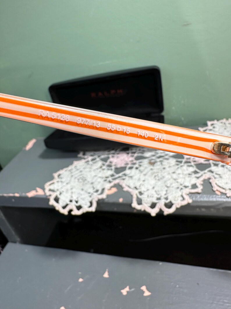 Puede incluir: Primer plano de la patilla de unas gafas de sol Ralph. La patilla es blanca con rayas naranjas y presenta el n&uacute;mero de modelo RA 5128, junto con otros n&uacute;meros. Un estuche negro para gafas est&aacute; en el fondo.
