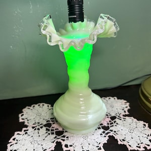 Könnte beinhalten: Eine hellgrüne Glasvase mit gerüschtem Rand, von innen mit einem hellgrünen Licht beleuchtet. Die Vase steht auf einer weißen Spitzendecke mit rosa Rändern. Eine schwarze Taschenlampe ist in die Oberseite der Vase eingesetzt.