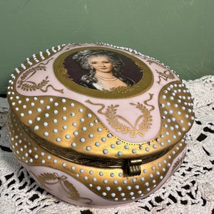 Puede incluir: Una caja de porcelana rosa y dorada con un retrato de una mujer en la tapa. La caja está decorada con puntos blancos y detalles dorados.
