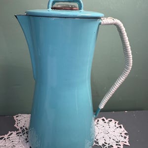 May include: A vintage turquoise enamelware coffee pot with a white wrapped handle and a glass-topped lid. The pot has a tall, slender shape and is likely from the mid-20th century. The handle is wrapped in a white material. The pot is sitting on a white lace doily.