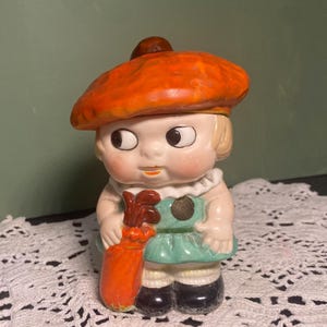 May include: A vintage ceramic figurine of a child wearing an orange beret and holding a golf club. The figurine has a green dress, white collar, and black shoes. The figure is standing on a white lace surface.