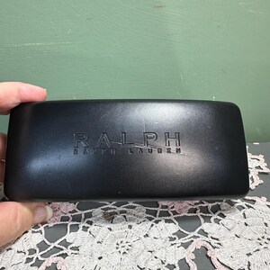 Puede incluir: Un estuche rectangular negro para gafas con el nombre de la marca "RALPH RALPH LAUREN" en relieve. El estuche est&aacute; sostenido por una mano y descansa sobre un tapete de encaje decorativo blanco y rosa.