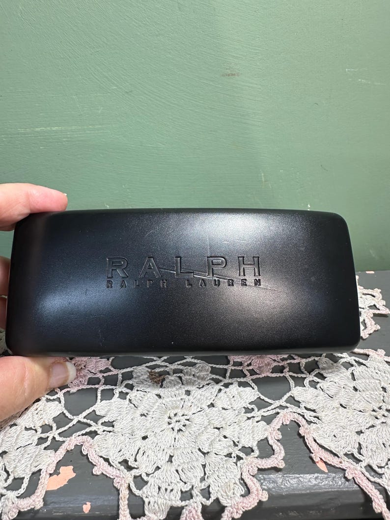 Puede incluir: Un estuche rectangular negro para gafas con el nombre de la marca "RALPH RALPH LAUREN" en relieve. El estuche est&aacute; sostenido por una mano y descansa sobre un tapete de encaje decorativo blanco y rosa.
