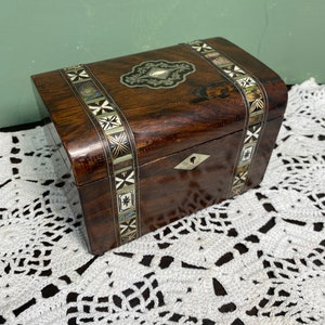 May include: A small wooden box with intricate inlaid designs. The box is made of dark wood and has a hinged lid. The lid is decorated with a silver-colored metal design and a small, round, white stone. The box is sitting on a white crocheted doily.