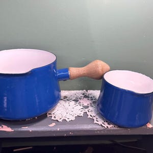 May include: Two blue enamelware pots with white interiors and wooden handles. The larger pot has a long handle, while the smaller one has a rounded handle. They are sitting on a gray surface with a white lace doily.