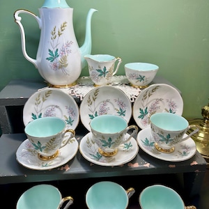 Puede incluir: Un juego de té vintage con una cafetera de porcelana blanca con tapa y asa azul claro, y tazas, platillos y una cremaera a juego. El juego está decorado con motivos florales en verde, rosa y detalles dorados.