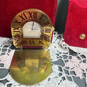 May include: A gold-toned Cartier clock with a rectangular face and Roman numerals. The clock has a hinged base and a red accent. The clock is next to a red velvet pouch.