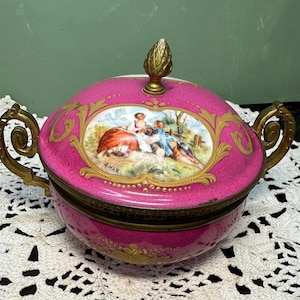 May include: A pink ceramic trinket box with a lid, featuring a gold-framed pastoral scene and ornate gold handles. The lid is topped with a gold finial. The box is set on a white lace surface.