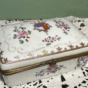 May include: A rectangular white porcelain trinket box with a hinged lid. The lid is decorated with floral patterns in pink, purple, and green, and a crest. The box has gold-colored metal trim and a clasp.