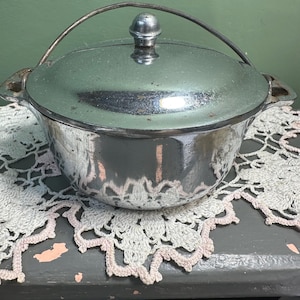 May include: A small, shiny, silver-colored metal pot with a lid and a handle. The pot has a rounded shape with two small handles on either side. The lid has a small knob on top. The pot is sitting on a decorative, white lace doily.