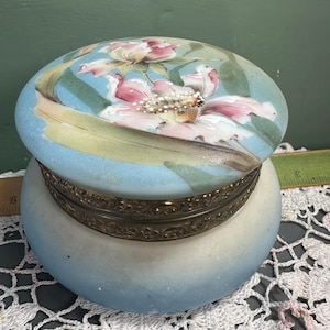 May include: A vintage, oval-shaped trinket box with a floral design on the lid. The lid features a light blue background with pink and white flowers and green leaves. The box has a brass-colored metal rim and a light blue and white body.