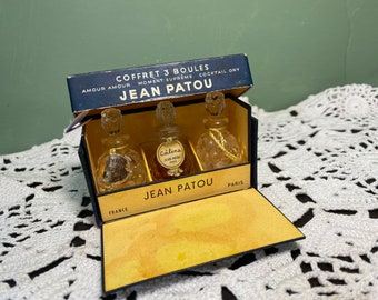Puede incluir: Una caja negra con interior dorado que contiene tres pequeños frascos de perfume de vidrio. La caja está etiquetada "Coffret 3 Boules Moment Supreme Amour Amour Cocktail Dry Jean Patou" y "Jean Patou Paris".