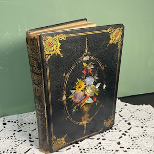 May include: Antique book with a black cover and gold accents. The cover features a floral arrangement in a gold oval frame. The book's spine has gold lettering. The book is resting on a white lace doily.