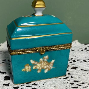 May include: A teal porcelain trinket box with a gold-colored rim and accents. The box features a floral design and a gold-colored knob on the lid. The box is rectangular with a hinged lid.