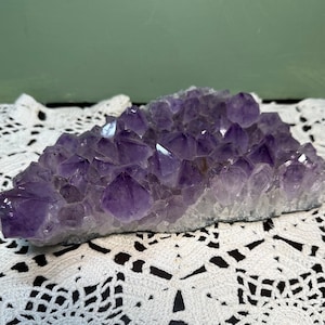 May include: A large amethyst crystal cluster with a purple and white color. The crystal is sitting on a white lace doily.
