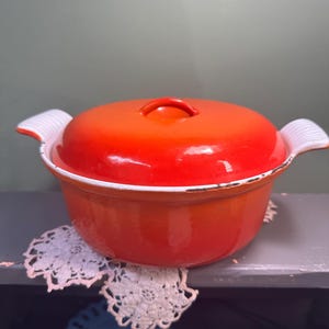 May include: A vibrant orange enameled cast iron Dutch oven with a matching lid. The pot has white handles and a white rim. The pot is sitting on a gray surface with a white lace doily.
