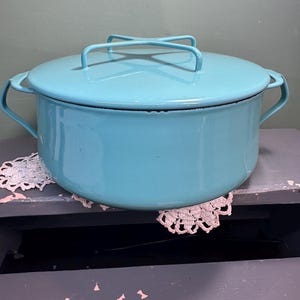May include: A light blue enamel cooking pot with a matching lid and two side handles. The lid has a cross-shaped handle. The pot is sitting on a distressed gray surface with a white lace doily.
