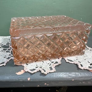 Könnte beinhalten: Vintage Schmuckkästchen aus rosa Glas mit Rautenmuster. Die rechteckige Box hat einen passenden Deckel und wird auf einer weißen Spitzendecke präsentiert. Ideal zur Aufbewahrung von Schmuck oder kleinen Andenken.