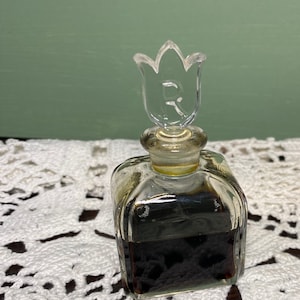 May include: A small, clear glass perfume bottle with a tulip-shaped stopper. The bottle is square with a dark liquid inside. The stopper has the letter "R" etched on it. The bottle sits on a white, lacy surface.