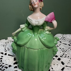 May include: A porcelain figurine of a woman in a green dress, holding a pink fan. The figurine has blonde hair with a pink flower and a white bodice. The base of the figurine is a box shape.
