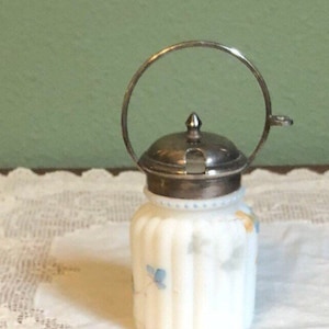 Könnte beinhalten: Eine weiße Milchglasdose mit einem silbernen Deckel und einem silbernen Ringgriff. Die Dose hat ein Blumenmuster mit blauen und gelben Blumen.