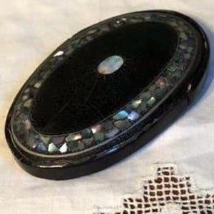 May include: Oval black box with a decorative mother-of-pearl border and a central iridescent stone. The box is set on a white, patterned surface, showcasing its intricate design and craftsmanship.