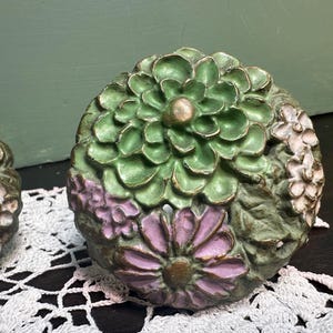 Könnte beinhalten: Zwei dekorative, blumenförmige Keramikknöpfe mit grünen, rosa und weißen Details. Die Knöpfe haben ein erhabenes, strukturiertes Design und sind perfekt, um Möbeln einen Hauch von Vintage-Charme zu verleihen.