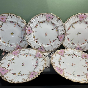 May include: A set of five white porcelain plates with a pink and gold floral design. The plates have a scalloped edge and are decorated with colorful flowers and gold trim. The plates are arranged on a dark surface.