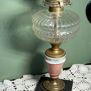 Könnte beinhalten: Eine antike Öllampe aus Glas mit einem klaren Glasschirm, einem rosa-weißen Porzellanfuß und einem Messingbrenner. Die Lampe steht auf einer weißen Häkeldeckchen.