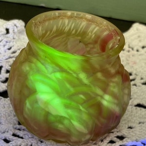 Pode incluir: Um pequeno vaso de vidro verde com um design floral. O vaso tem uma superfície texturizada e é feito de vidro de urânio, que brilha sob uma luz negra.