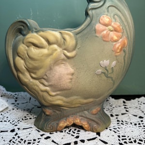 May include: A vintage art pottery vase in a muted green and peach color palette. The vase features a woman's profile and floral accents. The design has a crackle finish, adding to its antique aesthetic. The vase is approximately 10 inches tall.