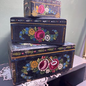 May include: Three stacked, vintage black boxes with hand-painted floral designs. The boxes feature pink, yellow, blue, and white flowers with gold and green accents. The largest box has a keyhole and a small key.