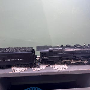 May include: Black model train with a coal car and engine. The coal car is labeled "NEW YORK CENTRAL." The engine has the number "5335" on the side. The train is sitting on a gray surface.