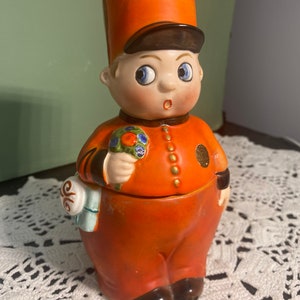 May include: A vintage ceramic figurine of a bellhop in an orange uniform and hat. The figurine has a surprised expression, blue eyes, and holds a bouquet of flowers. It has a small bag with a scroll design and gold buttons.