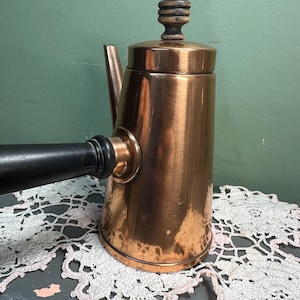 Może przedstawiać: Miedziany dzbanek do kawy z czarnym uchwytem i drewnianym pokrętłem na pokrywce. Dzbanek ma dzióbek i stoi na białej koronkowej serwetce. To przedmiot w stylu vintage.