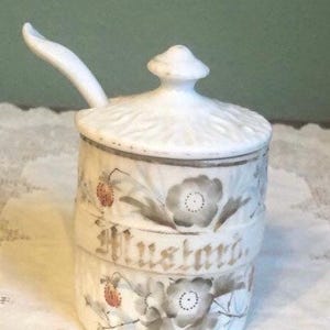 May include: White ceramic mustard jar with a lid and attached spoon. The jar is decorated with gray floral and strawberry designs, and the word "Mustard" is written in gold script across the front.