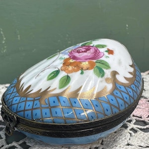 May include: An egg-shaped trinket box with a white top adorned with a floral design in pink, orange, and green. The base features a blue diamond pattern with gold accents. The box has a metal hinge and clasp.