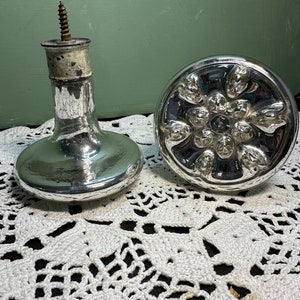 May include: Two silver glass bottle stoppers with a decorative design. One stopper has a screw top and the other has a round top with a floral design.