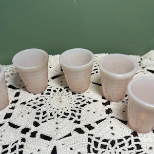 May include: Five small, pink, glass cups with a textured, waffle-like pattern. The cups are arranged in a row on a white crocheted doily.