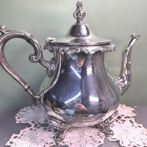 May include: A silver-colored teapot with ornate detailing, including a curved handle, a spout, and a decorative finial on the lid. The teapot sits on three small feet and reflects the surrounding environment.