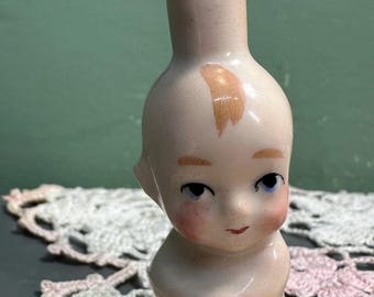 Może przedstawiać: Antyczna butelka w kształcie lalki z porcelany z mosiężnym korkiem w kształcie korony. Lalka ma bladą cerę, niebieskie oczy i różowe policzki. Na czole namalowany jest blond kosmyk włosów. Unikalny przedmiot dekoracyjny.