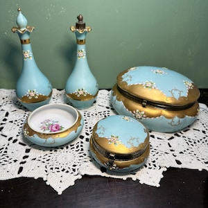 Puede incluir: Un conjunto de cinco artículos de tocador vintage de porcelana en un esquema de colores azul claro y dorado. El conjunto incluye dos botellas, un tazón pequeño y dos cajas redondas con tapas. La porcelana está decorada con delicados diseños florales en blanco y dorado.