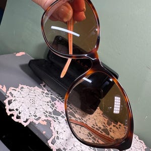 Puede incluir: Gafas de sol carey con lentes marrones. Las monturas tienen un dise&ntilde;o cl&aacute;sico de forma redondeada. Las patillas son de color naranja claro. Las gafas de sol se sostienen frente a un estuche negro.