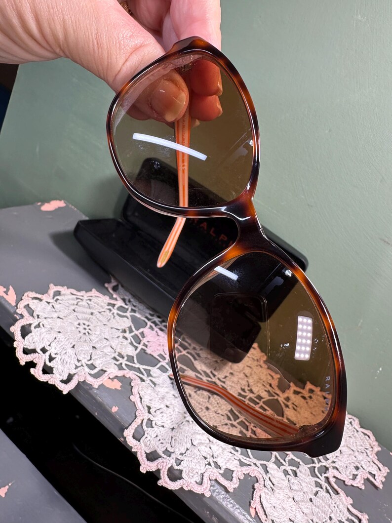 Puede incluir: Gafas de sol carey con lentes marrones. Las monturas tienen un dise&ntilde;o cl&aacute;sico de forma redondeada. Las patillas son de color naranja claro. Las gafas de sol se sostienen frente a un estuche negro.