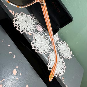 Puede incluir: Gafas de sol con montura a rayas naranja y blanca y lentes oscuras. Las gafas de sol est&aacute;n en un estuche negro con el nombre "RALPH" impreso. Las gafas de sol descansan sobre un tapete de encaje blanco.