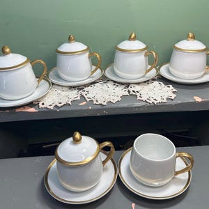 May include: A set of white porcelain teacups and saucers with gold detailing. Some cups have lids with gold finials. The handles and rims are also gold. The cups are arranged on a grey surface.